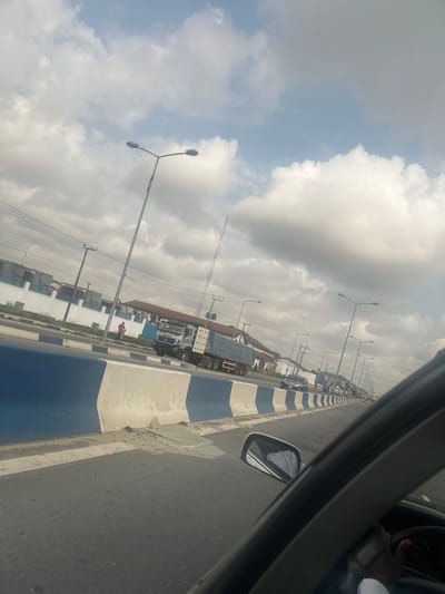 The image depicts a highway scene in Elimgbu, Nigeria, viewed from inside a vehicle looking forward and to the left. The sky is overcast with a mix of white and grey clouds.

In the foreground, the interior of a vehicle is visible, showing a portion of the dashboard, a steering wheel on the right, and the passenger side mirror. A person's arm, dressed in a white sleeve, is visible within the vehicle's interior.

The road features a paved surface and is separated by a concrete barrier painted alternately in blue and white. To the immediate left of this barrier is another lane of traffic. A large white truck with a blue tarp covering its rear section is positioned in this lane, appearing stationary or moving slowly. Further down this lane, additional vehicles are visible, forming a line.

To the left of the road, a narrow shoulder or sidewalk area runs alongside. A single individual, dressed in a red shirt, is visible walking on this area. Beyond the shoulder, a white wall with blue trim encloses a property with buildings and a brown-roofed structure. Numerous utility poles with overhead power lines and multiple streetlights with dual fixtures line the road. In the distance, a tall, slender antenna-like structure rises above the other buildings.