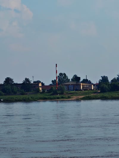 The image captures a wide, calm body of water in the foreground, reflecting the overcast sky. Across the water, a treelined bank is visible, behind which lies a cluster of industrial or commercial buildings. A prominent red and white striped smokestack stands tall among the structures. The buildings appear to be of varying sizes and designs, with some appearing older and weathered, and others newer. There are some vehicles visible near the buildings. The scene suggests a semi-industrial area adjacent to a river, possibly in the city of Yeniseysk, Russia, given the context provided. The sky is a pale blue with some scattered clouds.