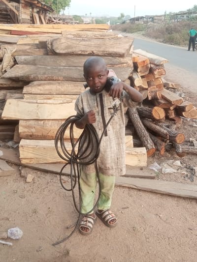 The image features a young child, appearing to be a boy, standing in an outdoor setting in Ebiya, Nigeria. The child is positioned centrally in the foreground, wearing a light-colored patterned tunic, light green trousers, and open-toed sandals. He holds a large coil of black cable in his left arm and has a black power tool, resembling an angle grinder or sander, resting on his right shoulder/back. He looks directly at the camera with a neutral expression.
Behind the child, substantial stacks of raw logs and roughly sawn timber planks dominate the scene. To the child's right, additional cut logs are neatly piled. In the background, a dirt road is visible, on which a man in a green shirt and dark trousers walks away from the camera. Further beyond the road, there are hints of simple wooden structures and sparse vegetation under what appears to be a clear, bright sky. The lighting suggests it is daytime, likely late morning or afternoon, given the soft illumination and lack of harsh shadows. There is no visible text in the image. The scene conveys a working environment, possibly a timber market or processing area.