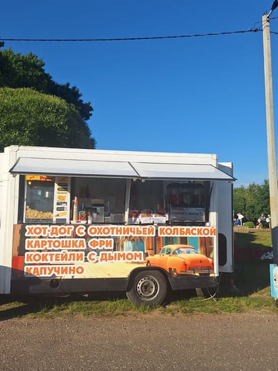 A food truck is parked on a grassy area under a bright blue sky with some trees in the background. The food truck is white with a large graphic on its side featuring an orange classic car and text in Russian. The text advertises various food items like hot dogs, french fries, cocktails, and cappuccino. Inside the truck, there are visible signs with prices and product names. People are seen in the background, some sitting on the grass and others standing, suggesting a public outdoor setting, likely in the vicinity of Цімкавіцкі сельскі Савет, Belarus. The overall scene appears to be a sunny day with a relaxed atmosphere.