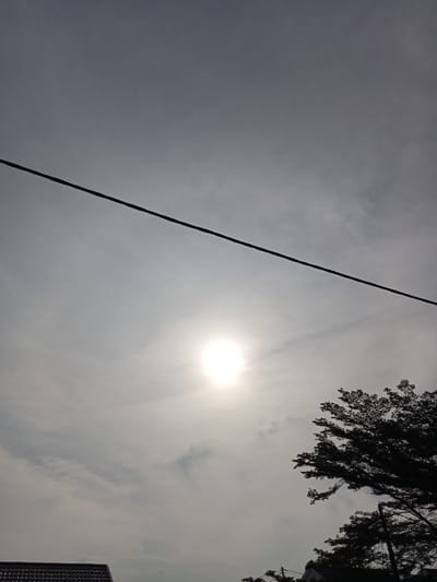 The image captures a bright sun partially obscured by hazy clouds, positioned in the upper center of the frame. A dark, diagonal power line stretches across the upper portion of the image, from the left edge towards the right. In the lower right quadrant, the silhouette of a large tree with dense foliage is visible against the bright sky. The lower left corner shows a sliver of a dark-tiled rooftop, likely belonging to a house. The environment suggests an outdoor scene in the city of Kualu, Indonesia, likely during daylight hours given the sun's prominence. There are no people or discernible actions taking place in the scene.