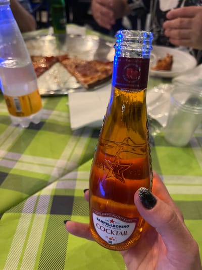 A close-up shot shows a person's hand holding a small glass bottle of San Pellegrino Cocktail. The bottle is amber-colored and has a red label with white text. The hand has black nail polish with glitter on one nail. In the background, out of focus, is a table covered with a green and white checkered tablecloth. There are slices of pizza on a metal platter, a plastic water bottle, and several disposable cups. The overall scene suggests a casual meal, possibly a pizza party. The image does not provide any specific information about the location within Toritto, Italy.