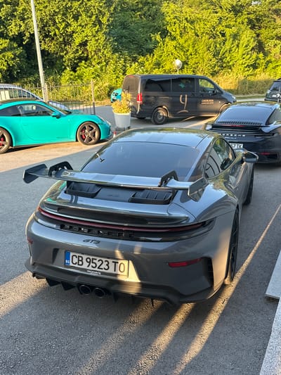 The image depicts an outdoor scene with several cars parked on an asphalt surface. The main focus is a grey Porsche GT3 with a large rear spoiler. To its left is a bright teal Porsche. Behind the grey Porsche, a black Mercedes-Benz van is visible with the logo "CHUKARA WINE & SPA HOTEL" on its side. Further to the right, another black Porsche is parked. The environment is characterized by lush green trees and foliage in the background, suggesting a location with abundant vegetation. Sunlight casts long shadows across the ground, indicating it is likely late afternoon or early morning. The license plate of the grey Porsche is visible and reads "CB 9523 TO", with "BG" indicating it is a Bulgarian registration. The website "www.porsche.bg" is also printed below the license plate. The scene appears to be a parking area, possibly at a hotel or a resort, given the presence of the wine and spa hotel van. The location is identified as Kerenite, Bulgaria. There are no people visible in the image.
