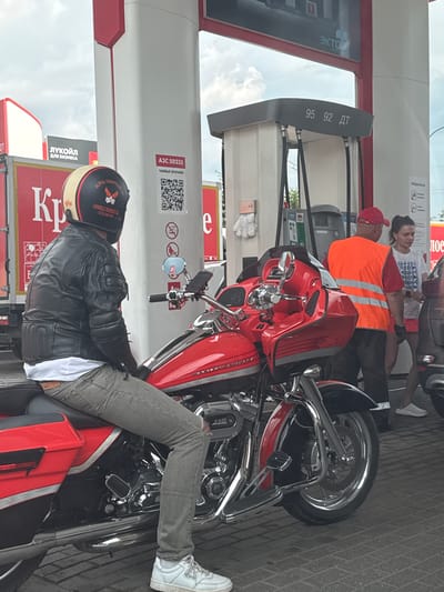 In Ermolino, Russia, at a gas station, a person is seated on a red motorcycle. The individual is wearing a black leather jacket, grey jeans, and white sneakers. A helmet with a white, yellow, and black design is on their head. The motorcycle is positioned in front of a gas pump. Behind the motorcycle, a gas station attendant in an orange safety vest and a woman in a white t-shirt and pink shorts are visible. The environment appears to be outdoors under an overcast sky, with gas pumps and advertising signage present.