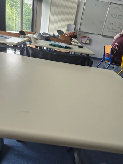 The image depicts an indoor setting, likely a classroom or office space, in Thiais, France. The focus is on a table, which dominates the foreground and appears to be the surface of a desk or conference table. The table is light beige and clean. To the left of the table, one can see a row of tables with personal items, including a laptop, notebooks, and a bag.
In the background, a whiteboard is visible on the left wall, featuring handwritten notes in French. There is a person partially visible in the upper portion of the image wearing a maroon shirt and sitting in a yellow and blue chair. The lighting is bright, suggesting natural light streaming through a window. The room also has an air of functionality and order, indicating it's a place for work, study, or meeting. The presence of personal items on the tables suggests that it's a space used by individuals.