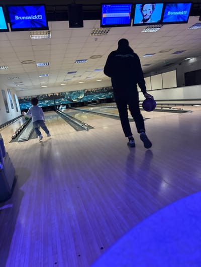 The scene is inside a bowling alley. An individual is captured in mid-swing, preparing to release a bowling ball onto the lane. Nearby, another person is visible at the end of the lane. A series of screens display bowling scores and advertisements for Brunswick bowling equipment. The environment suggests an indoor recreational setting. The photo was taken in Gioia Tauro, Italy.