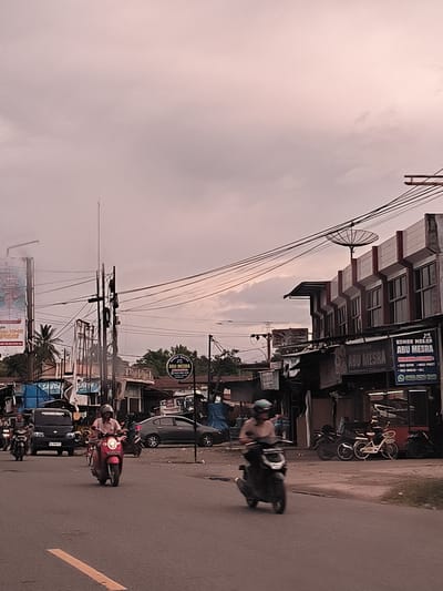 This image captures a bustling street scene in Grong-Grong, Indonesia, likely during the late afternoon or early evening under an overcast sky. The diffused, warm light casts a muted, somewhat desaturated tone over the entire scene.

The foreground features a paved road with a yellow lane marker, where several motorbikes are in motion. A person wearing a helmet and light-colored shirt is riding a red scooter directly towards the viewer in the mid-ground, while another person on a dark scooter is partially blurred on the right, indicating movement. A black van is visible behind the red scooter, and another motorbike and a dark sedan are further back on the street.

The sides of the street are lined with low-rise commercial buildings, some with awnings and storefronts. Overhead, a dense network of electrical and communication wires crisscrosses the sky, connected to numerous utility poles. A large billboard with partially visible text is on the left, next to a palm tree. On the right, a building features a prominent satellite dish on its roof.

Visible text includes multiple signs for "ABU MESRA," indicating a business, with one sign explicitly stating "RUMAH MAKAN ABU MESRA" (Abu Mesra Restaurant) and further details like "MENERIMA PESANAN" (Accepts Orders), "RENTAL MOBIL & BUS" (Car & Bus Rental), and a phone number "0822-5250 7845." Another blue tarp or sign reads "SAUDA." Partial text on the billboard includes words like "SUMBING," "BOCOR," and "PIGIE." "PRUPAKALAY" is also visible on a sign.

The scene conveys the everyday rhythm of a busy street in an Indonesian town, characterized by diverse vehicles, numerous overhead wires, and local commercial establishments.