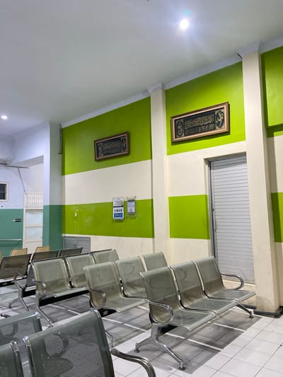 This media file shows a waiting area, likely in a public building or institution, characterized by its green and white striped walls and tiled floor. The scene is dominated by rows of metal chairs, suggesting a place designed to accommodate multiple people waiting for an appointment or service. The chairs are modern, with a mesh-like material for the seat and back, and chrome accents.
On the walls, there are two framed pieces of calligraphy, likely religious texts, adding a cultural or spiritual element to the environment. Above the chairs, a horizontal window with vertical blinds is visible. There are also some signs on the wall, one of which appears to display information with icons, possibly related to health or safety guidelines. Another sign clearly indicates "TOILET" with an icon.
The lighting suggests it is daytime, with a prominent light fixture visible on the white ceiling. The overall impression is one of a clean, functional, and organized space, typical of a waiting room. The image is taken from a low angle, emphasizing the rows of chairs and the striped pattern of the walls. The scene appears quiet and empty, with no people present, implying the waiting area is currently unoccupied. The location context provided is Margoyoso, Indonesia.