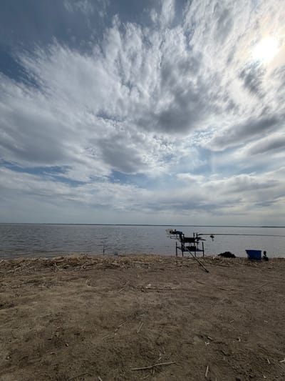 The image shows a lakeside view on what appears to be a cloudy day near Krasnyi Bor, Russia. A body of water (possibly a lake or river) occupies the middle ground, with a shoreline on the right. On the shoreline, there's a fishing setup which includes fishing rods, a fishing rod stand, and a blue container. The foreground shows dry, earthen ground with some dried vegetation. In the sky, clouds dominate the view. The sun is visible behind some clouds.