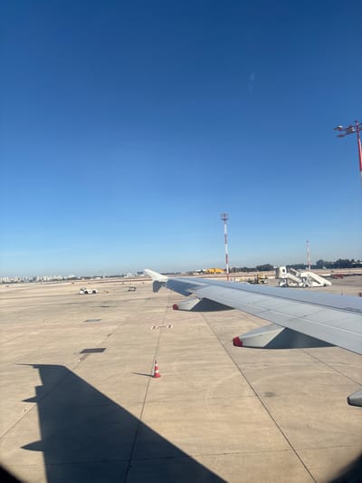 An airplane wing is visible in the upper right of the image, casting a shadow on the concrete runway. In the background are airport support vehicles and a tall red and white communications tower. The sky is a clear, gradient blue, taking up the majority of the view. Red and white traffic cones are positioned on the runway.