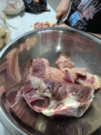 An overhead shot displays a large, reflective stainless steel bowl filled with raw meat. The meat appears to be various cuts, with some pieces exhibiting a reddish-brown hue and others being lighter, possibly containing fat or poultry. The bowl is positioned on a white countertop with a subtle marble pattern. To the left, a clear plastic bag contains what looks like round, pale food items, possibly bread or dumplings. In the background, a white bowl with a white cloth draped over it, and a black plastic bag containing more food items are visible. A person's hands, wearing a black t-shirt with graphics, are visible to the right, holding a knife and in the process of preparing the meat. The scene is likely in a kitchen environment.