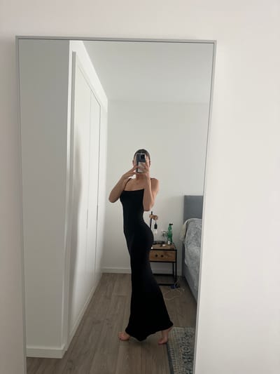 An individual is depicted taking a mirror selfie in an indoor room located in Ericeira, Portugal. The person is standing barefoot, wearing a form-fitting black sleeveless maxi dress with spaghetti straps. They are positioned facing slightly to the left, with their right arm raised, holding a smartphone with a light-colored case that obscures their face, capturing their reflection in a large, rectangular full-length mirror. The mirror has a thin, light-colored frame. The environment reflected in the mirror includes a modern bedroom setting. To the left, a white built-in wardrobe with multiple flat-panel doors is visible. To the right, behind the individual, a wooden bedside table with two drawers supports a dark-colored lamp featuring an exposed bulb and a green bottle. A bed with a grey upholstered headboard and a grey and white patterned duvet is partially visible behind the bedside table. The room features plain white walls and light brown wooden plank flooring. A small, patterned rug lies on the floor near the bed.