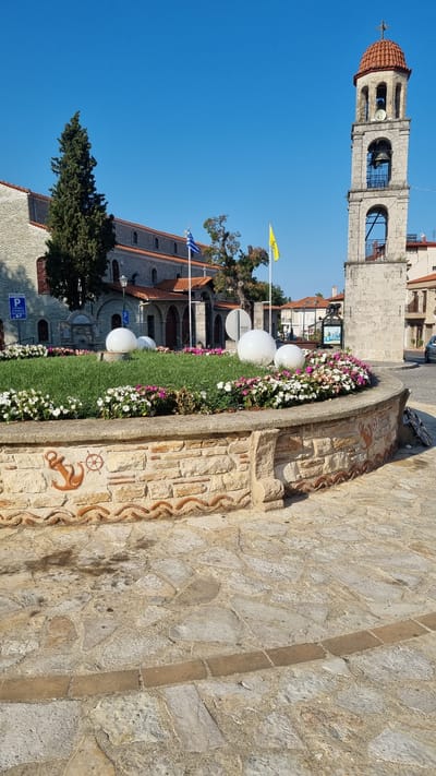 This image captures a public space in Litochoro, Greece, featuring a stone-paved area in the foreground with a circular flowerbed at its center. The flowerbed is bordered by a low, multi-textured stone wall adorned with decorative elements, including an anchor and a ship's wheel motif. Inside the flowerbed, vibrant green grass is interspersed with blooming flowers in shades of pink and white, and two large, white spherical objects are placed strategically.
In the background, a multi-tiered bell tower with a red tiled roof stands prominently. Adjacent to the tower is a stone building with an arched entrance, likely a church, indicated by a cross at the apex of its roof. The Greek flag and a yellow flag are displayed near the buildings. Several trees, including a tall cypress tree, are visible, adding to the greenery of the scene. The sky above is a clear, bright blue, suggesting fair weather. A street with parked cars is visible to the right, and a signpost with Greek text is present near the left side of the frame, indicating a parking area. The overall impression is of a well-maintained public square with architectural and natural elements, typical of a Greek town.
