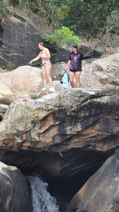 Two women stand on a rock formation near a waterfall in what appears to be a natural outdoor setting. One woman wears a bikini and points toward the waterfall. The second woman wears a dark t-shirt, shorts, and glasses. The scene is dominated by rocks, foliage, and the flowing water of the waterfall. The location is Nam Ninh Hoa, Vietnam.