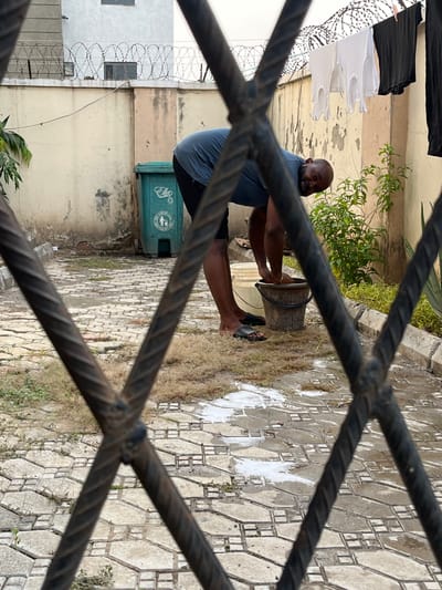 This image, captured in Dushepe, Nigeria, portrays a man engaged in outdoor domestic work, viewed through the prominent, dark, twisted bars of a metal grate or fence. The scene is presented with the man vertically oriented, suggesting a 90-degree rotation from the original capture.
The primary subject is a dark-skinned man with a shaved head and a beard, wearing a dark blue t-shirt and dark shorts. He is barefoot, with a black sandal visible near his left foot. He is focused on a task, with both hands submerged inside a dark, aged bucket resting on the paved ground in front of him, suggesting he is washing or soaking items. White, foamy residue, likely from soap or detergent, is visible on the ground around the bucket.
In the background to the man's left, several white and black sleeveless shirts are hanging on a clothesline, indicating laundry is taking place. Beyond the clothesline, a wall or fence topped with multiple strands of barbed wire runs horizontally. Further back, weathered building walls in shades of light tan and off-white are visible, along with some green foliage.
To the man's right and slightly behind him, a green plastic waste bin with a lid is visible. Text on the bin includes "KEEP CLEAN" and a logo depicting a hand dropping an item into a bin. The ground is paved with patterned concrete or interlocking tiles, interspersed with patches of dry grass and dirt. The time of day appears to be daylight, under natural illumination.