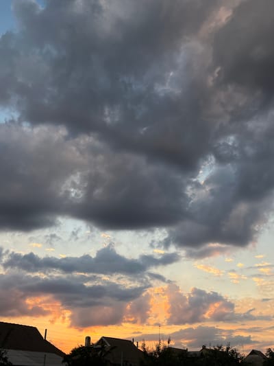 A wide-angle shot captures a dramatic sky over rooftops in Slobozia, Moldova. The upper portion of the frame is dominated by dark, ominous storm clouds, with hints of lighter blue sky peeking through in some areas. Below these, the clouds transition to a more vibrant sunset, with swathes of orange and yellow light illuminating puffy cloud formations. The lower edge of the image shows the dark silhouettes of several rooftops, some with visible chimneys, and the tops of trees. There are no people or discernable actions visible in the scene. The overall impression is of an approaching storm at dusk.