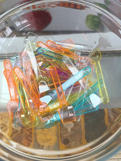 The image shows a close-up, overhead view of a pile of small, colorful plastic scoops or spoons, predominantly orange, yellow, pink, and blue, jumbled together in a clear glass bowl. The scoops are scattered and overlapping, with their handles and scoop heads visible. The bowl itself has a metallic sheen and is transparent, revealing a blurred background that appears to be a surface with a wood-grain pattern. Reflections of what seem to be objects or light sources are visible on the surface of the bowl. The scene is brightly lit, causing some glare and highlighting the translucent nature of the scoops. There are no people visible in the image. The location in the city of Águilas, Spain, is not depicted or implied by the visual elements.