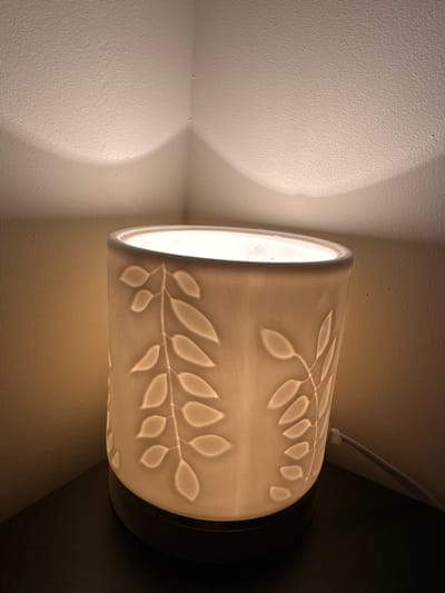 An illuminated, cylindrical lamp with a leaf pattern is positioned on a dark surface, likely a nightstand or table, in a corner of a room. The lamp's light casts soft shadows on the textured walls behind it. A grey power cord is visible, connected to the lamp. The scene is indoors, with no specific landmarks to identify a location within Cumming, United States.