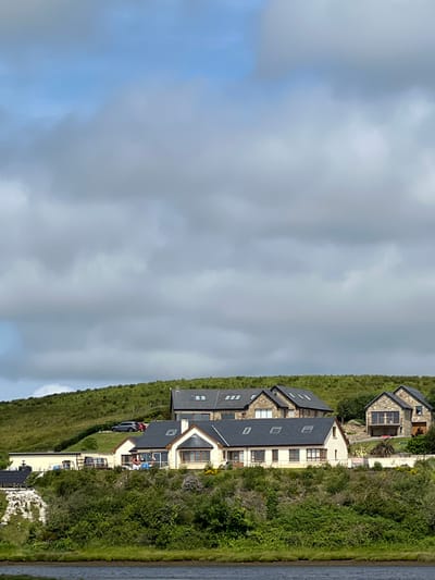 The image shows a group of modern houses situated on a grassy hillside overlooking a body of water. The architecture of the houses features stone facades and dark tiled roofs. Some houses have large windows and balconies. The surrounding landscape is characterized by rolling green hills covered in dense vegetation, with patches of rough terrain. The sky above is partly cloudy with a mix of blue sky and grey clouds. A few cars are parked on the upper part of the hillside. The foreground shows the edge of the water, with a strip of dark soil or vegetation along the bank. The scene appears to be a residential area in a rural or coastal setting.
