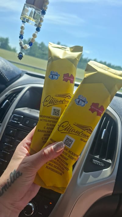This media file captures a close-up view of a person's hand holding two yellow ice cream wrappers inside a car. The wrappers feature text in Cyrillic script, including "Коммунарка" (Kommunarka) and "Столичное" (Stolichnoye). One wrapper also displays "МОРОЖЕНОЕ" (ice cream). The hand, with a sparkly manicure and a tattoo on the wrist, is holding the wrappers at an angle, obscuring parts of the car's interior, which appears to be a dashboard with air vents and control buttons. To the left, a car accessory, possibly a car air freshener adorned with beads and a hamsa charm, hangs from the rearview mirror. The background shows a bright, sunny sky and blurred greenery, suggesting it's daytime with good weather. The overall scene implies a casual moment, perhaps during a road trip or a pause in a journey. The product name "Столичное" (Stolichnoye) and the brand "Коммунарка" (Kommunarka) suggest that the ice cream is likely from Belarus, given the location context.