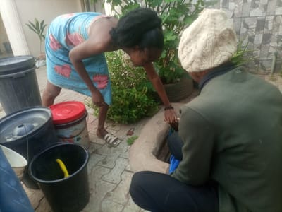 In Wuro Dole, Nigeria, a scene unfolds involving two individuals amidst various containers and greenery. A person wearing a blue and pink patterned wrap bends over, reaching towards a collection of black and red plastic buckets. Their posture suggests they are engaged in an activity involving the contents or arrangement of these containers. Next to them, another individual, seen from the back, wears a green hooded garment and a cream-colored knit hat. They are seated or crouching near a large, curved object that appears to be made of concrete or earth. The environment is paved with textured tiles, and lush green plants are visible, some growing in what look like repurposed tires. The overall setting appears to be an outdoor domestic or communal space.