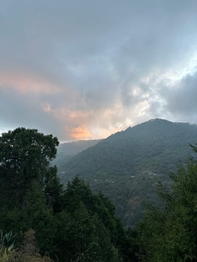A mountain looms with dense green foliage on its steep slope. The landscape is shrouded in overcast skies, with hints of golden light breaking through the clouds near the mountain's peak. Trees frame the lower right corner of the image, adding depth to the view. The overall atmosphere is tranquil and somewhat misty.