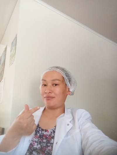 A woman in a white lab coat and hair net smiles at the camera, holding up her hand with her thumb and index finger touching to form a small heart shape. She is wearing a floral patterned top underneath the lab coat. The background is a plain white wall with some framed pictures hanging on it to the left. The lighting suggests it is daytime. The woman appears to be in a casual, perhaps work-related setting. The images on the wall are not clear enough to read any text. The overall emotion conveyed is friendly and approachable.