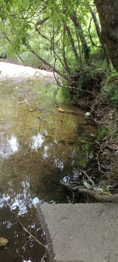 The image depicts a shallow creek flowing through a wooded area. The water is clear enough to see the rocky bottom. Debris, including a white plastic ball and what appears to be a yellow plastic bag, is floating in the water near the right bank. The creek banks are lined with trees and vegetation. A concrete structure is visible in the foreground.

While I can describe the scene, I do not have information to identify the specific location as being in Tahlequah, United States of America.