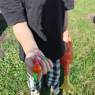 In Podlubovo, Russia, a person wearing a black t-shirt and plaid pants stands in a grassy area, holding a cherry tomato on a skewer. The person's nails are decorated with colorful designs, including polka dots and a watermelon pattern. To the right, an orange watering can is visible. The scene appears to be outdoors, likely in a garden or backyard.