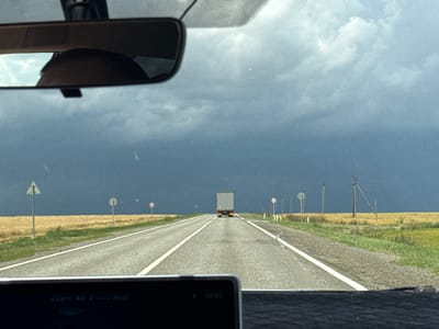 This media file depicts a view from the inside of a vehicle, looking out through the windshield and a rearview mirror. The scene is a road journey through a rural landscape under a dramatic sky.
The primary subjects are the road, a truck in the distance, fields of dry grass or crops lining the road, and the sky above. A rearview mirror shows a reflection of a person's head. The road is paved and marked with white lines, indicating it's a highway. Several road signs are visible along the roadside, including a triangular sign with a white background and a red border, and circular signs with red borders. In the far distance, a large grey truck is traveling in the same direction. The landscape consists of flat fields of golden-brown grass or harvested crops to the left and right of the road, with scattered green vegetation closer to the road.
The activity is clearly a car journey, suggesting travel. The weather appears to be a significant element, with a dark, stormy sky dominating the upper half of the image. The contrast between the dark, brooding clouds and the brighter, sunlit fields creates a sense of dramatic weather. The light suggests it could be late afternoon or early evening, given the potential for storm clouds to gather during that time.
Notable details include the reflection of the driver in the rearview mirror, hinting at the presence of people. The road signs are subtle but provide context for a road environment. The distinct dark storm clouds are the most striking feature, suggesting impending rain or a storm. There is text visible on a small screen at the bottom of the image, which appears to be a car display showing temperature and time. Partially visible text includes "23.0°C" and "20.0°C", and "13:33".
The setting is a rural road in Svetlograd, Russia, characterized by open fields and a vast sky. The overall mood conveyed is one of travel through a dynamic and potentially turbulent weather system.