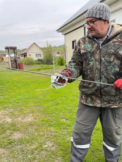 A middle-aged man wearing a gray knitted hat, glasses, a camouflage fleece jacket, and gray work pants with reflective stripes is standing in a grassy area, holding a metal rod with a cloth attached to one end. He is wearing red gloves. The setting appears to be a rural or semi-rural location, with a house visible in the background, along with a metal structure that could be related to water heating or plumbing. A second house is visible further in the background. The sky is overcast. The location is in Talmenka, Russia.