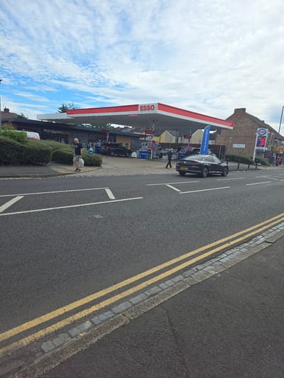 The image shows a street scene in Stockton-on-Tees, United Kingdom. In the foreground, there is a road with double yellow lines and a pavement. In the background, there is a petrol station with an Esso sign. A man is walking on the pavement carrying shopping bags. A black car is driving on the road. There are trees and buildings in the background, and the sky is blue with some clouds.