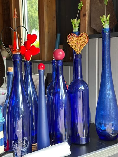 The image depicts an indoor scene in Deinste, Germany, showcasing approximately ten cobalt blue glass bottles of various shapes and sizes, arranged on a black, reflective surface. Several bottles are fitted with decorative stoppers or inserts: a translucent orange, pointed, helical stopper; four red fabric hearts on individual sticks; a small red wooden spherical stopper; a yellow wire-wrapped heart; and two translucent green plastic stoppers shaped like stylized flowers. A natural cork stopper is also visible in one bottle. One bottle displays a white printed label with text identifying it as "1995ER RIVANER QUALITÄTSWEIN TROCKEN RHEINHESSEN GUTSABFÜLLUNG." An opaque, matte blue, conical object is positioned among the other bottles. The background features a window revealing green foliage, vertical wooden planks, a dark metal heart-shaped stand, and white vertical wall paneling. A white ceramic bowl with a rose pattern and a white plastic item are present in the lower-left background and foreground, respectively. No human figures are depicted.