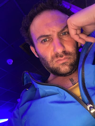 The image is an inverted, close-up selfie of a man in an indoor setting, illuminated by dominant dark blue and purple artificial lighting. The man, likely in his 30s or 40s, has a dark, full beard, dark hair, and blue eyes. He looks directly at the camera with a neutral expression. His left hand is raised, partially resting on his forehead or hair.
He is wearing a bright blue zippered jacket over a yellow shirt, visible at the collar. On his neck, a tattoo is partially visible, appearing to read "YNNEN" in capital letters. The background consists of a dark wall or ceiling with a subtle geometric pattern. A white, circular object, possibly a smoke detector or sensor, is visible on the dark surface in the lower right portion of the original image, along with a dark air vent. The blue and purple lighting creates a cool, atmospheric ambiance, suggesting an evening or nighttime setting, possibly in a public entertainment venue or a room with mood lighting. No specific visual cues within the image indicate the location as Armagh, United Kingdom, beyond the provided context.