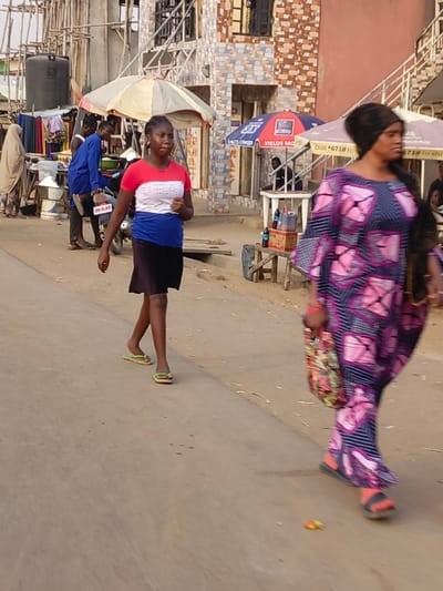 The image captures a vibrant street scene, likely in a market or commercial area, during daytime. The lighting is bright but diffused, with no harsh shadows, suggesting it could be late morning, early afternoon, or an overcast day.
In the foreground, two women are walking on a light-colored, unpaved road. On the left, a young woman with dark skin and braided hair is walking towards the viewer's left. She is wearing a short-sleeved t-shirt with horizontal stripes of red, white, and blue, and a black skirt that reaches her knees. She has light green flip-flops on her feet. Her expression is neutral as she looks slightly ahead. On the right side of the frame, partially cut off, another woman with dark skin is also walking towards the viewer's left. Her head is covered with a dark fabric, and she is wearing a long, loose-fitting dress or gown with a distinctive geometric pattern in shades of blue and pink/purple. She carries a floral-patterned bag in her left hand and wears dark sandals with an orange/red strap. Her face is slightly blurred, indicating movement.
In the midground, behind the young woman, there is a bustling street with various stalls and people. To the left, under a light-colored, possibly white, umbrella, a vendor is present. A person dressed in light-colored clothing and a head covering is visible near a white bucket or container, suggesting a stall selling goods. Clothes are hanging on a line behind this stall. A man in a blue top is leaning on or near a dark-colored motorcycle parked on the side of the road. Part of a license plate or sign on the motorcycle is visible, showing "GB HOL".
Further back on the right, under a blue and red umbrella, another vendor stall is set up. Text visible on this umbrella includes "AITSTORMER" and "YIELDS MONEY". A person can be seen sitting behind a table under this umbrella. On a small wooden stand in front of this stall, several plastic bottles, possibly containing drinks or cooking oils, are displayed for sale. Another umbrella, further in the background, also has visible text, including "Dial *671#" and "Dial *673#".
The background features multi-story buildings, some appearing to be under construction or renovation, with wooden scaffolding visible. The walls of these buildings are clad with patterned tiles in shades of brown, grey, and orange, adding texture to the scene. The overall impression is one of everyday life in a lively street environment.