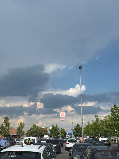 The image depicts a drive-thru lane at a Burger King in Singen (Hohentwiel), Germany, on a partly cloudy day. The lane is filled with numerous cars, suggesting a busy period. A Burger King sign with the iconic crown logo is visible, along with a "Drive Thru" sign. Trees line the perimeter of the area, and streetlights are present. In the foreground, a white car is parked, and to its left, a man in a blue shirt stands near another vehicle. A woman in a black and white patterned dress is seen standing near a white van with lettering on its rear. The sky is a mix of blue and dark, dramatic clouds, indicating potential impending weather changes.