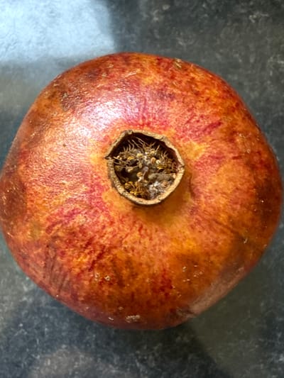 A close-up, overhead shot shows a ripe pomegranate resting on a dark, speckled surface. The pomegranate is a vibrant reddish-orange, with subtle striations of darker red and yellow. Its skin appears slightly textured, with a few small, lighter speckles visible. At the top of the fruit, a small, dark, crown-like protrusion is visible, with dried remnants of the flower still attached. The background surface is a dark gray, possibly granite or a similar countertop material, showing a slight sheen. There are no people or discernible actions in the image. The scene is static and focuses solely on the pomegranate. The location of Drumettaz-Clarafond, France, is not depicted or implied by the image content.