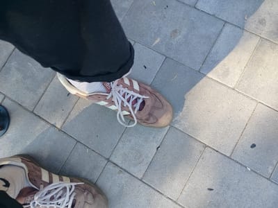 The image shows a close-up view of two Adidas sneakers on a grey paved surface. The shoes are a muted pinkish-brown color with the characteristic white Adidas stripes. The laces are white and loosely tied. One shoe is more prominent in the foreground, with the wearer's black pants and white socks visible. The other shoe is visible in the lower left corner of the image, partially obscured. The paving stones are rectangular and laid in a slightly staggered pattern. There are some small shadows cast on the ground, suggesting it is daytime, possibly late afternoon due to the warm light. The overall setting appears to be outdoors, in a public area like a street or park. The image is taken from a high-angle perspective, looking down at the shoes and ground. There is no discernible text in the visible portion of the image, beyond the Adidas branding on the shoes themselves. The scene is casual and everyday, with no specific event or activity being depicted, other than walking or standing.