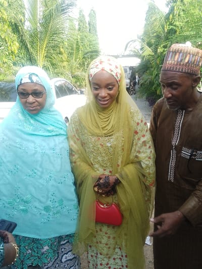 The image depicts three individuals standing outdoors in Unguwa Uku Kauyen Alu, Nigeria, under bright daylight. On the left, an older woman with dark-framed glasses wears a light blue hijab over a white patterned band and a matching embroidered garment, partially revealing a dark patterned lower garment. A hand holding a dark-colored phone is visible at the bottom left. In the center, a younger woman with a slight smile wears a light green embroidered dress with red floral patterns, a sheer green veil over a white and red patterned head covering, and displays intricate black henna designs and multiple gold rings on her right hand, which holds a small, bright red rigid handbag with a gold handle. On the right, an older man wears a brown traditional tunic with white chest embroidery and a multi-colored patterned cap, looking downwards and to his left. The background consists of dense green foliage, including palm trees, with a white vehicle partially visible behind the left woman. Other distant vehicles and structures are faintly discernible.