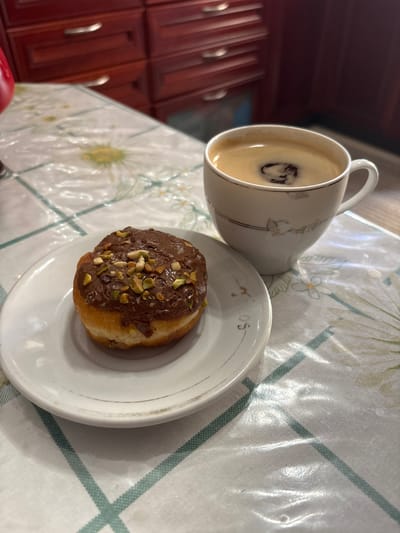 A close-up, eye-level shot shows a single donut on a white plate and a white coffee cup filled with dark coffee on a table covered with a patterned tablecloth. The donut, positioned on the left side of the frame, is round with a hole in the center, covered in a dark chocolate glaze and sprinkled with chopped pistachios. The white plate has a delicate gold trim. The coffee cup, to the right of the donut, is also white with a gold trim and contains coffee with a frothy surface. In the background, dark red wooden cabinets are visible, suggesting a kitchen or dining area. The image appears to be taken indoors. The question about the location in Ilion, Greece, cannot be answered from the provided image.