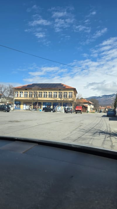 The image depicts a street scene in Slatino, Bulgaria, viewed from inside a vehicle. A large, four-story yellow building with solar panels on its roof is visible on the left side of the street. The building appears to have retail or commercial spaces on the ground floor. Cars are parked along the street. In the background, there are mountains. The sky is blue with scattered clouds. The foreground is dominated by the interior of the vehicle.