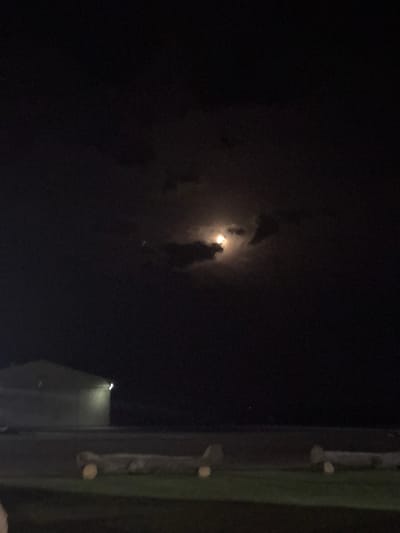 The image depicts a nighttime scene in Port Alfred, South Africa. The upper two-thirds of the frame are dominated by a dark sky, featuring a bright, full moon partially obscured by dark clouds. A faint, singular light source is visible in the sky to the left of the moon. In the midground, a light-colored, corrugated metal building with a sloped roof is visible on the left, illuminated by a bright, white light fixture mounted on its upper exterior. The foreground consists of dark ground cover, likely grass, where two elongated, log-like structures are positioned horizontally. Each structure is supported by two lighter-colored, circular objects, resembling wheels or props. No individuals are present in the scene, and no discernible actions or interactions are occurring.