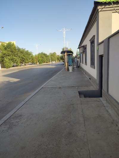 The image depicts a street scene in Ashgabat, Turkmenistan, under a clear blue sky. A paved road runs diagonally across the frame, bordered by a concrete sidewalk on the right. The sidewalk features a bus stop shelter with a trash receptacle. Adjacent to the sidewalk is a building with a light-colored facade, grey trim, and a green corrugated metal roof. Several windows with dark bars are visible on the building. Trees with green foliage line the opposite side of the road. In the distance, a few vehicles are present on the road, and a street lamp stands near the bus stop. The ground of the sidewalk is cracked and shows some wear. There is a visible step leading down from the sidewalk into a darker area next to the building. No individuals are prominently featured or interacting.