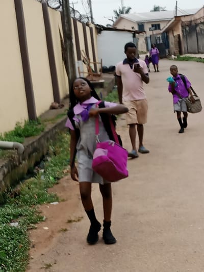 The image depicts an outdoor street scene in what appears to be a residential area in Crescent, Nigeria, under an overcast sky. In the foreground, a young girl, likely a student, walks along an unpaved or lightly paved road. She wears a grey uniform dress or overall with a purple shirt and bow tie, black socks, and black shoes. She carries a black backpack and a vibrant magenta shoulder bag, and her gaze is directed upwards.
Behind her, in the middle ground, a man in a light pink polo shirt and khaki shorts walks, engrossed in his phone. Further down the path, a young boy in a purple shirt and grey shorts carries a patterned shoulder bag and appears to be holding a light blue item. Two additional figures, also dressed in purple, are visible in the distance, suggesting they might be part of the same group or also going to or from school.
To the left, a tall, light-colored wall with prominent brown vertical stripes is topped with coiled barbed wire. Green vegetation borders a ditch or drain running alongside the path. Residential buildings are visible in the background. The children's school uniforms suggest the activity is a daily commute, likely to or from school. The time of day appears to be daytime, with soft, diffused lighting due to the cloudy weather. No visible text is present.