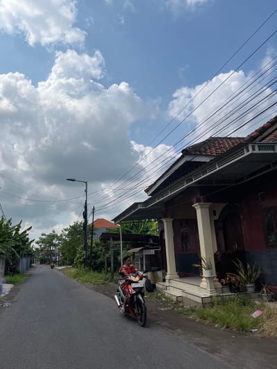 A motorcyclist, a woman wearing a red floral-patterned dress, is traveling on a paved road in what appears to be a residential area. She is on a red and black motorcycle with the license plate W 5399 DA. She is looking forward and appears to be alone on the motorcycle. In the background, there is a building with a dark red facade, white columns, and a tiled roof. There are also power lines visible against a blue sky with scattered white clouds. The area appears to be a street in Jati Alun Alun, East Java. The environment is a mix of residential buildings and vegetation.