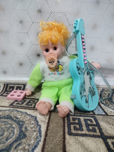 A doll with blonde hair and a pacifier sits on a patterned rug next to a blue toy guitar. The doll is wearing a green and white outfit with the words "Tough Pear" printed on the front. To the left of the doll is a pink wooden block with four circular indentations. The background is a white textured wall with a geometric pattern. This scene is located in Cholpon, Kyrgyzstan.