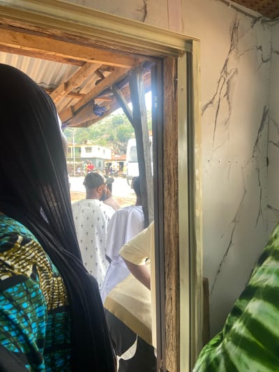 The image, taken from a low, interior perspective, looks out from a partially constructed structure towards a populated area in Okengwe-Obehira, Nigeria. The interior view reveals a frame made of wooden planks and beams, possibly part of a building under construction or renovation. Outside, a cluster of buildings and streets suggests a village or town, with people visible in the distance, indicating general activity. A vehicle can also be seen. The image is angled, and the perspective is such that it appears as though the viewer is looking out from within this incomplete structure, towards a public space. Parts of people or things are also in the shot.