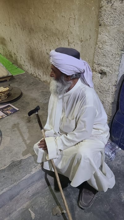 The image shows an elderly man in Ziarat, Pakistan, lying on the floor next to a wall. He is wearing traditional white clothing and a turban. He appears to be resting with a walking stick nearby. There is a round object, possibly a traditional cooking tool, and a newspaper on the floor next to him. The floor and wall show signs of age and wear.