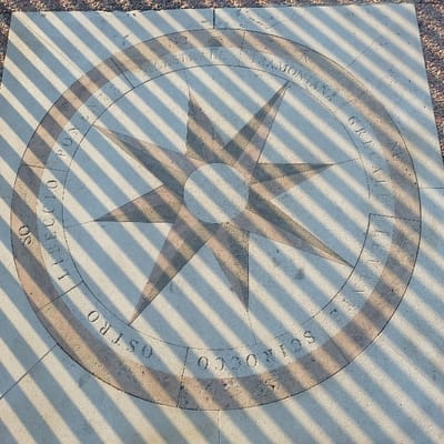 The image displays a large, circular stone compass rose inlay, approximately two meters in diameter, embedded in a light-colored, textured paved ground surface. This installation is located on the Lungomare Giacomo Matteotti in Pescara, Italy. The compass rose design features an eight-pointed star at its center, composed of alternating lighter and darker, possibly reddish-brown, stone segments. An outer circular band is inscribed with the Italian names of the eight principal winds and their corresponding directions: "TRAMONTANA" (North), "GRECALE" (Northeast), "LEVANTE" (East), "SCIROCCO" (Southeast), "OSTRO" (South), "LIBECCIO" (Southwest), "PONENTE" (West), and "MAESTRALE" (Northwest). No people are visible. The scene is illuminated by direct sunlight, casting numerous distinct, parallel diagonal shadows from an unseen overhead structure across the entire compass rose and the surrounding pavement.
