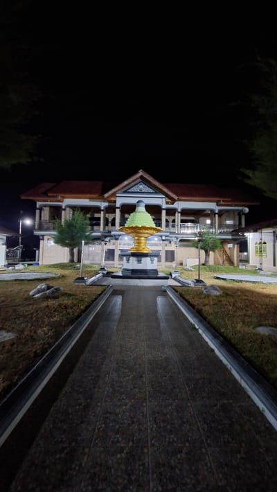 This is a nighttime, eye-level, full shot of a building in Lhokdagang, Indonesia. The building is two stories tall with a red, sloped roof and a white facade. The ground floor is a light brown brick, and the second floor features a balcony with white railings and visible columns. A golden, tiered fountain with a green top is situated in the foreground, centered on a paved walkway that leads to the building. The walkway is flanked by dried grass and scattered rocks. Trees are visible on the left and right sides of the frame, and the sky is completely black. The scene is illuminated by artificial lighting, creating a stark contrast between the brightly lit building and fountain and the dark surroundings.